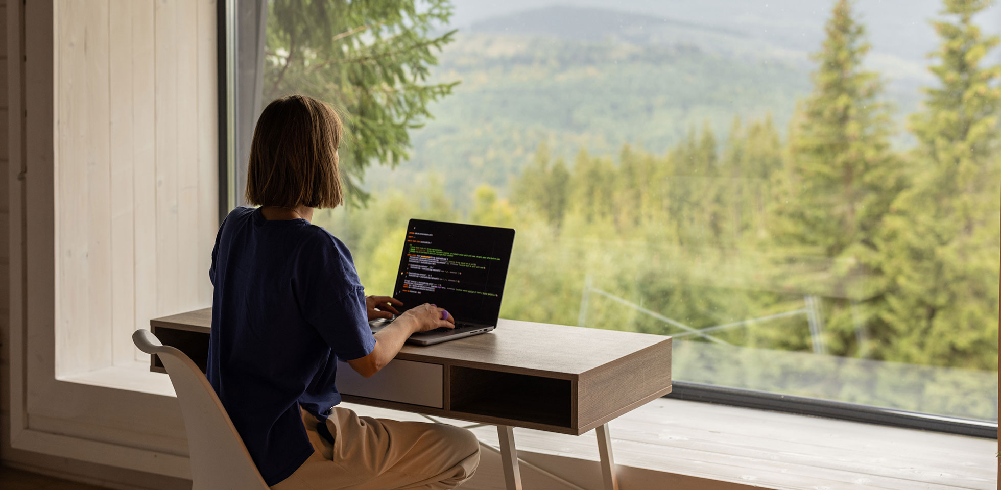 Une femme travaille sur son ordinateur portable, assise à une table devant une fenêtre panoramique offrant une vue imprenable sur les montagnes. Concept de travail à distance et d'évasion dans la nature.